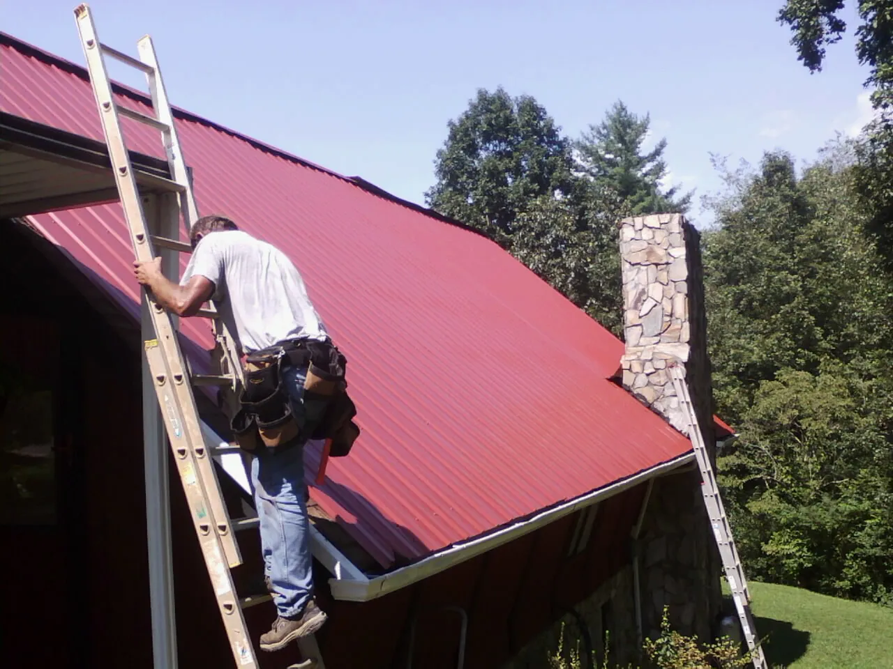 Roofer on a ladder beside a red metal roof and stone chimney — Soffit & Fascia Repair by Roofing Brothers in Medfield